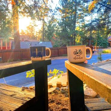 Two decorated mugs placed on chairs at a cabin - the sunshine is beaming through the trees and leaves, onto the mugs.