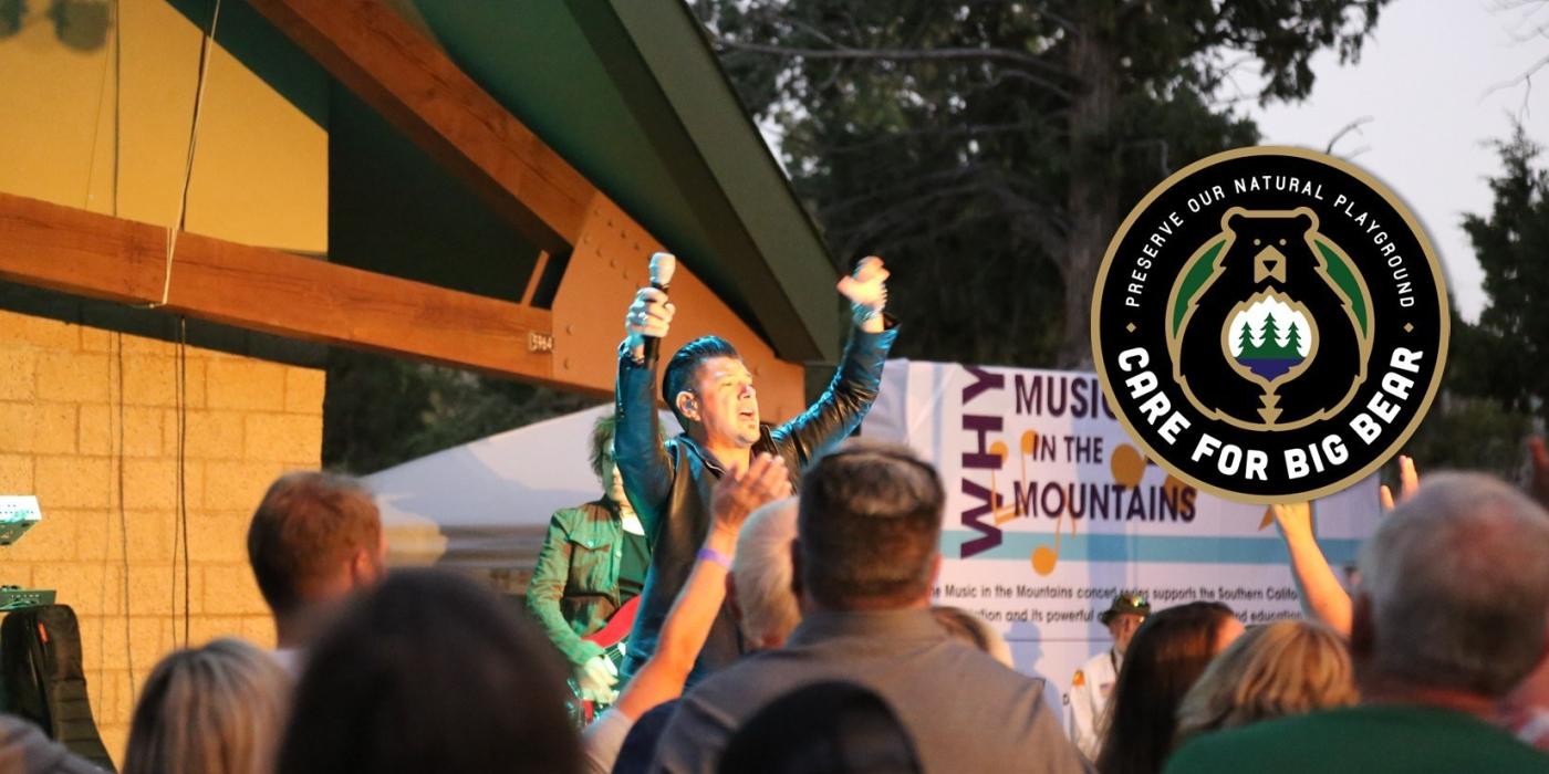 A man standing up with his arms raised at the Music In The Mountains concert, the care for big bear logo is displayed.