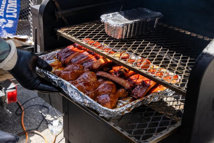 A tray of saucy barbecued meats being pulled out from a smoker - there are two metal racks on the inside of the smoker.