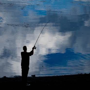 A silhouette of an angler stands on the shoreline of Big Bear Lake and casts a fishing line into the water. The water is blue
