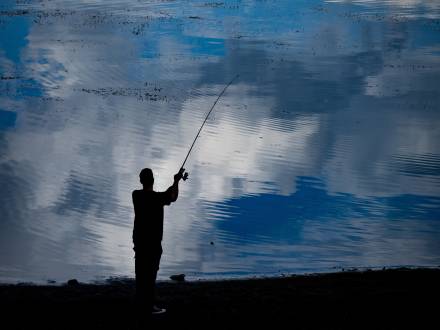 A silhouette of an angler stands on the shoreline of Big Bear Lake and casts a fishing line into the water. The water is blue