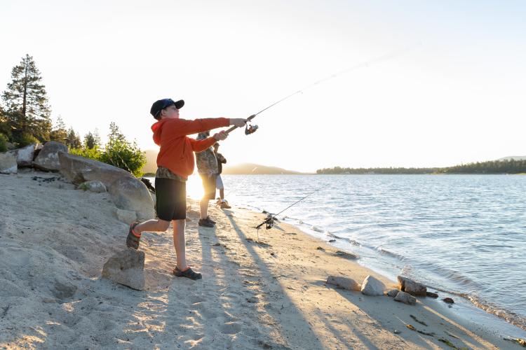 A boy in a red jacket casts a fishing line into Big Bear Lake while fishing on the beach with his family