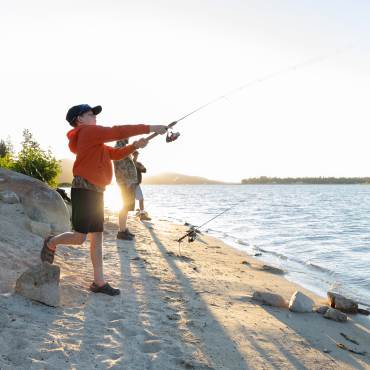 A boy in a red jacket casts a fishing line into Big Bear Lake while fishing on the beach with his family