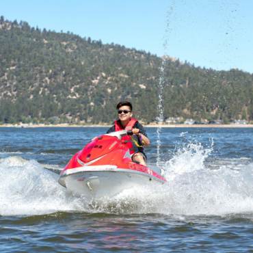 A person in a life jacket rides a red jet ski across a body of water, creating waves in the summer in  Big Bear Lake, CA.
