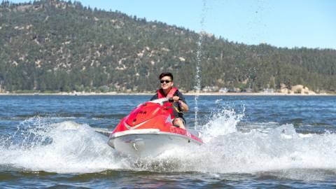 A person in a life jacket rides a red jet ski across a body of water, creating waves in the summer in Big Bear Lake, CA.