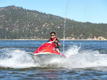 A person in a life jacket rides a red jet ski across a body of water, creating waves in the summer in  Big Bear Lake, CA.