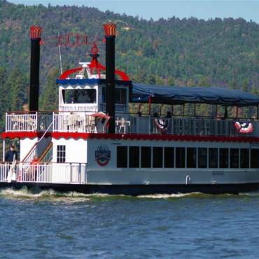 The two-story Miss Liberty Paddlewheel boat cruises along Big Bear Lake on a sunny day. She is painted red, white, and blue.