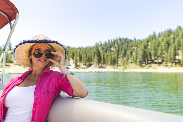 A female on a pontoon boat - she is holding her sun glasses looking out of view, the lake and trees visible behind her.