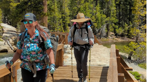 A female and male taking a hiking trail onto a bridge, they have hiking sticks and backpacks .