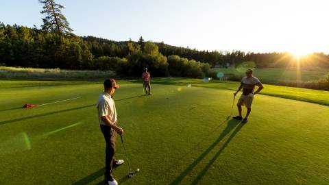 Three golfers standing on the lush green readying to putt at Bear Mountain Golf Course - the sun, mountains, and trees behind