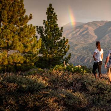 A photo of two people hiking on a trail in Big Bear California - there is a rainbow in front of mountains in the distance.