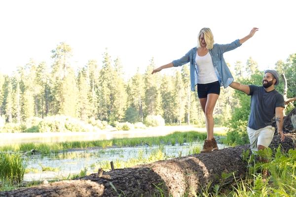 A photo of a couple outdoors with a girl being supported by a guy as she walks over a log.