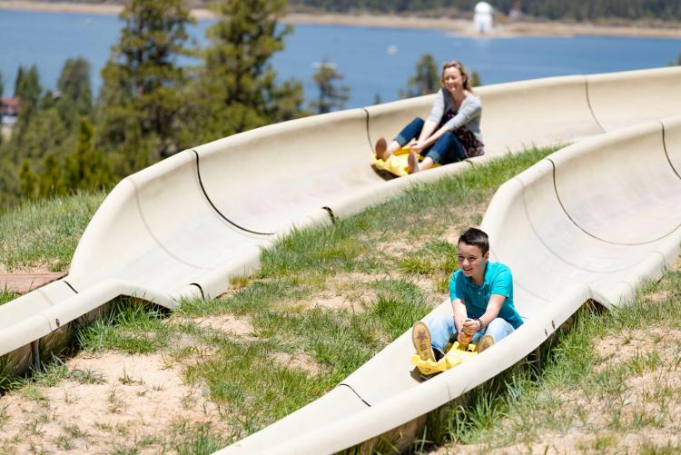 A mother and son riding on the Alpine Slide using yellow slide carts, the lake and solar observatory visible in the distance.