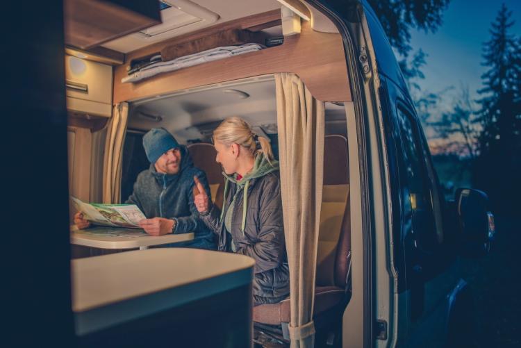 A couple chats inside a camping RV in a surrounding forest.