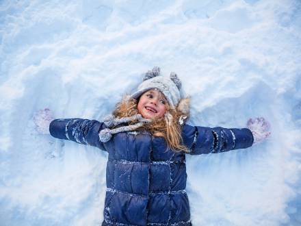 A photo of a little girl in a blue snow jacket making an angel in the fluffy white snow while smiling happily.