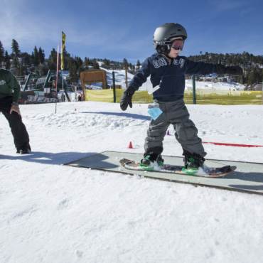 A young snowboarder slides across a rail during a lesson. His instructor looks on.