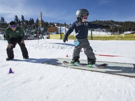 A young snowboarder slides across a rail during a lesson. His instructor looks on.