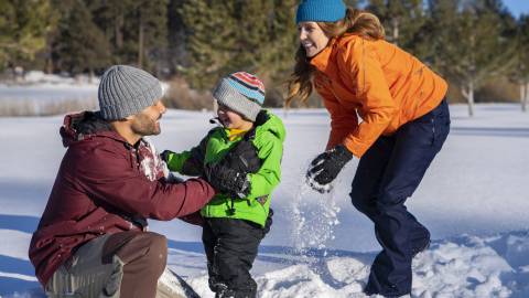 A mom and dad play with their son in Big Bear during the winter season. Snow covers the ground, and they are all smiling.