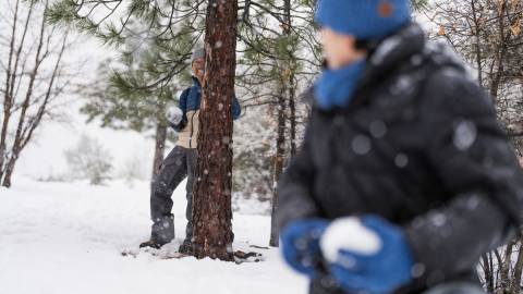 A photo of two people holding snow balls in the snow - one of them is hiding behind a tree while smiling.