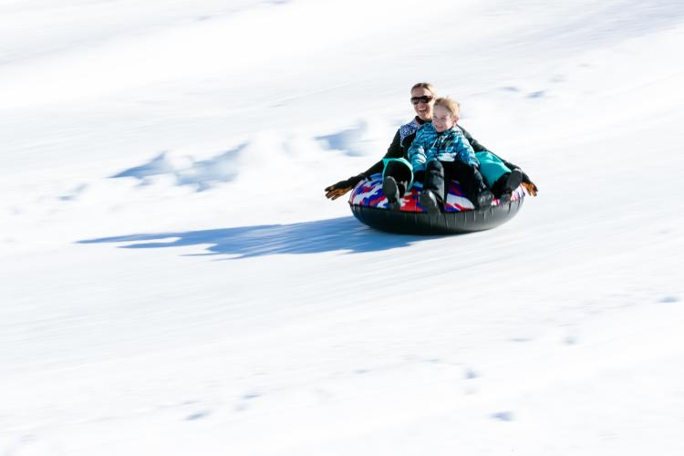A photo of a mother and son racing down a snow covered hill riding an inner tube, they are warmly bundled and smiling.