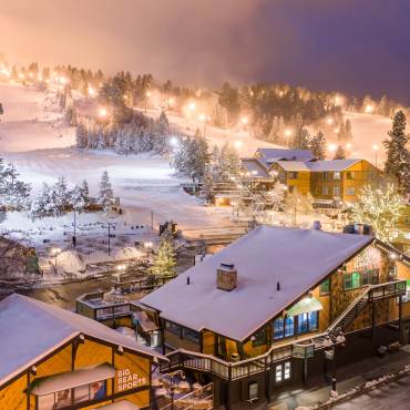 Snow Summit is illuminated at night for night skiing sessions during the winter season. Snow covers the buildings and trees.