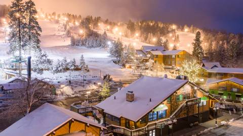 Snow Summit is illuminated at night for night skiing sessions during the winter season. Snow covers the buildings and trees.