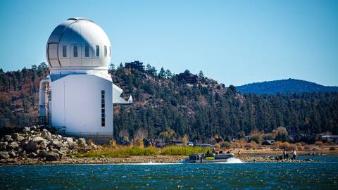 The white observatory dome stands tall over the Big Bear Lake shoreline. A pontoon boat sails by.