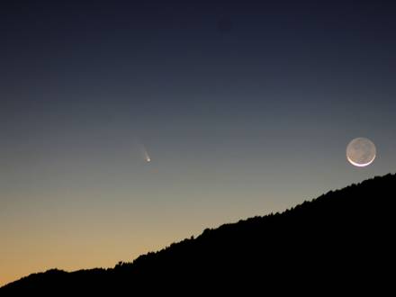 A photo of a silhouetted mountain, the moon is in a waning crescent stage as a comet is passing far away.