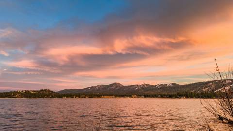 A shoreline view of Big Bear Lake - the lake below as well as the clouds above are gently glowing with a pink-orange sunset.
