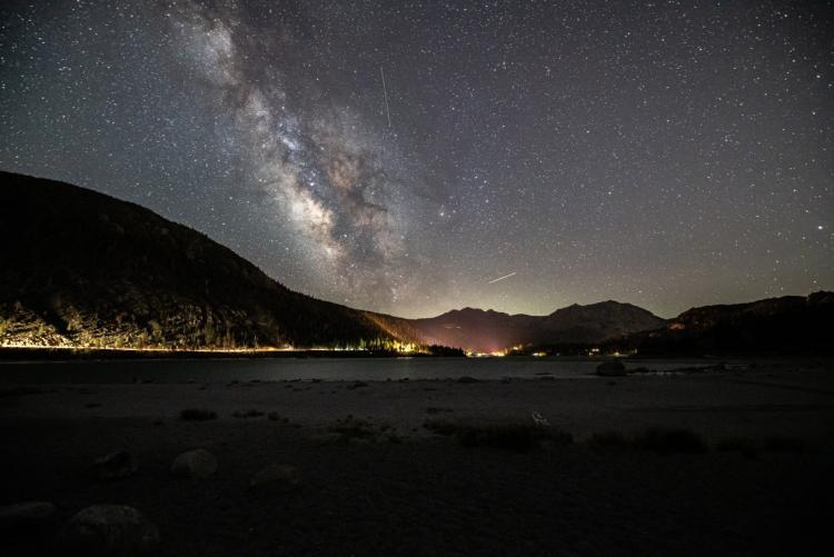 A photo of Big Bear's Night Sky - the milky way galaxy along with many stars are visible above the gently lit mountains.