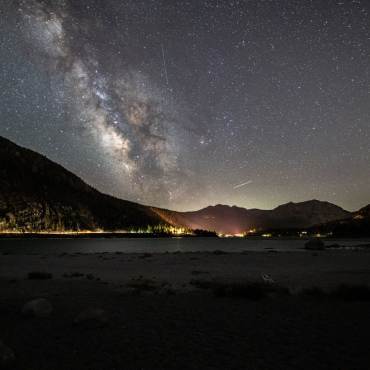 A photo of Big Bear's Night Sky - the milky way galaxy along with many stars are visible above the gently lit mountains.
