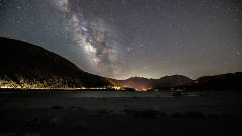 A photo of Big Bear's Night Sky - the milky way galaxy along with many stars are visible above the gently lit mountains.