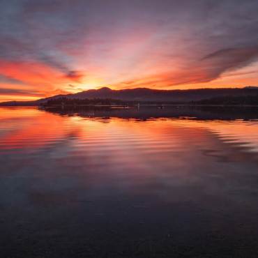 A photo of a bright red-ish pink and orange sunset peering from over the tall mountains, the lake reflecting it like a mirror