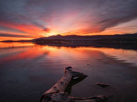A photo of a bright red-ish pink and orange sunset peering from over the tall mountains, the lake reflecting it like a mirror