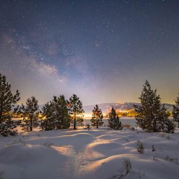 A photo of Big Bear's night sky taken during winter after a snowfall, the bright stars and a galaxy are visible.