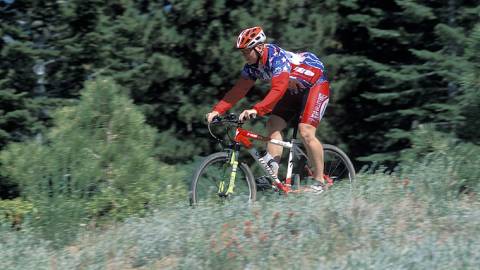 A mountain biker in a bright red kit rides along a bike trail in Big Bear Lake, CA. The lush, green forest surrounds him.