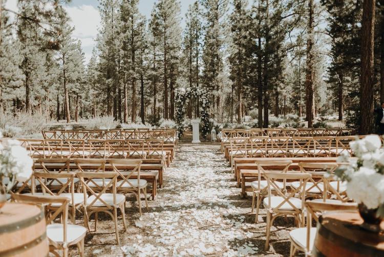 Wooden chairs sit in rows at a forested wedding venue in Big Bear. White flowers sits on wine barrels in the foreground.