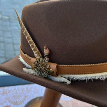 A custom hat made by Lucky Baldwin's - the leather brown colored hat is decorated with a feather sticking up on the side.
