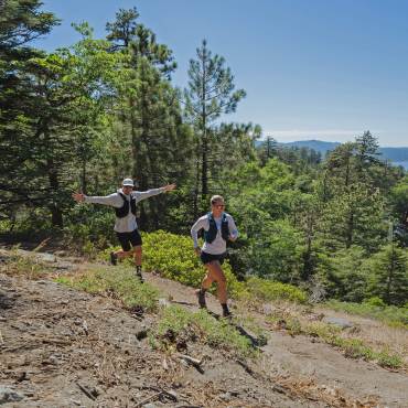 A photo of two hikers walking on a dirt hill trail - the dense forest and mountains are visible behind them.
