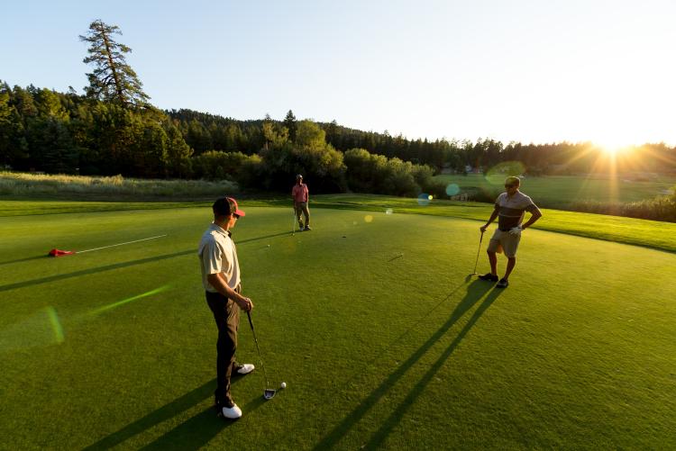 A group of three males golfing at Bear Mountain Golf Course on a flat green field - the trees and bright sunshine are visible