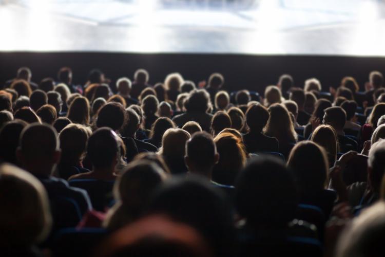 A photo of large movie theatre with an audience watching something on the widescreen in front of them.