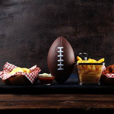 A football sits on a wooden table with appetizers including french fries and chicken wings. Stock image.