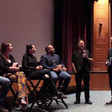 A group of film directors sitting in chairs listening to a presenter speak into a microphone during a Film Festival.