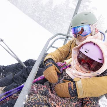 A photo of two people holding skis on a chair lift at Snow Summit Ski Resort in Big Bear Lake - snow is falling around them.