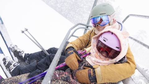 A photo of two people holding skis on a chair lift at Snow Summit Ski Resort in Big Bear Lake - snow is falling around them.