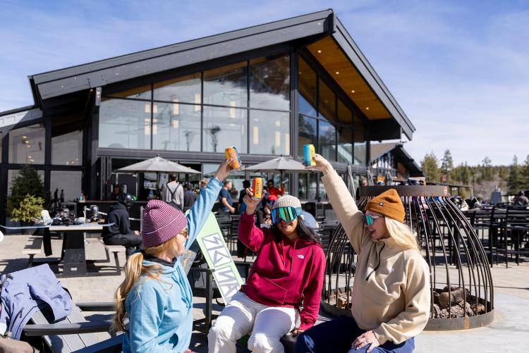 A group of three women wearing ski/snowboard equipment, raising up cans of Athletic Brewing Non-Alcoholic Beer.