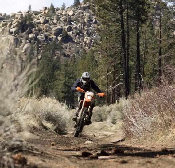 A dirt bike rider is racing down a dirt path on his orange bike, behind him is a rocky mountains and forestry.