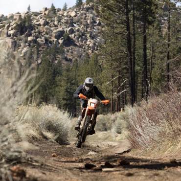 A dirt bike rider is racing down a dirt path on his orange bike, behind him is a rocky mountains and forestry.