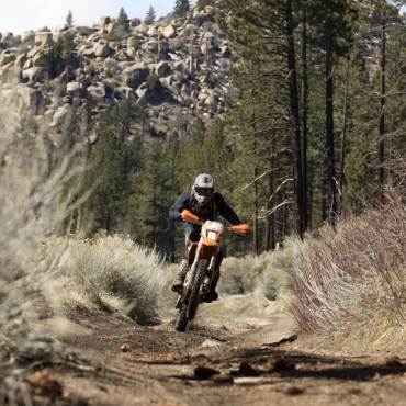 A dirt bike rider is racing down a dirt path on his orange bike, behind him is a rocky mountains and forestry.