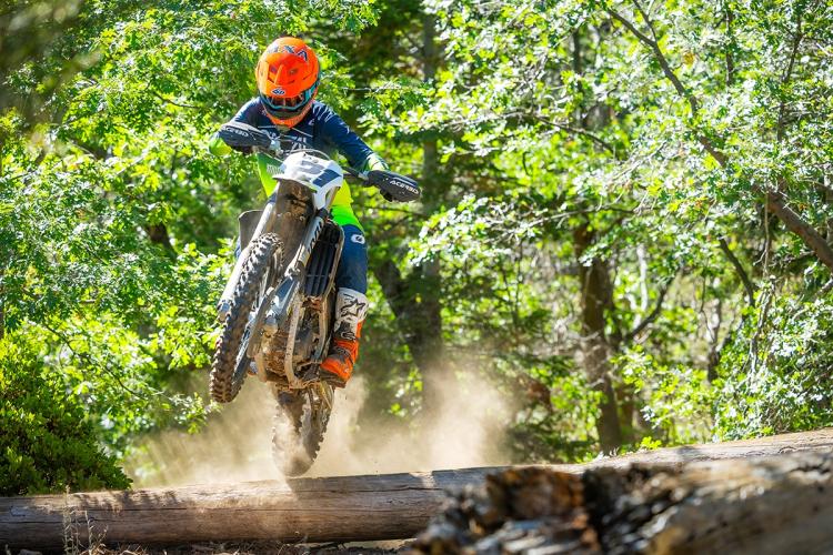 An MX-Bike rider seen mid-air on their bike while performing a jump over a log in a forest - a cloud of dust is behind them.
